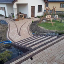 Ukończony podjazd i ścieżka z kostki brukowej dwukolorowej prowadząca do domu z drewnianą huśtawką w ogrodzie, widoczne schody z kostki. Niebieski wąż leży na ziemi obok.