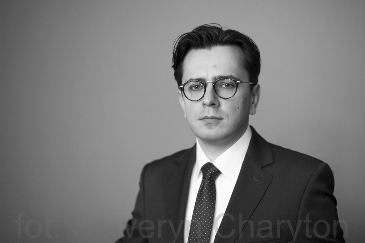 Portret biznesowy mężczyzny w garniturze i okularach, czarno-białe zdjęcie z subtelnym logo fotografa w dolnej części kadru.