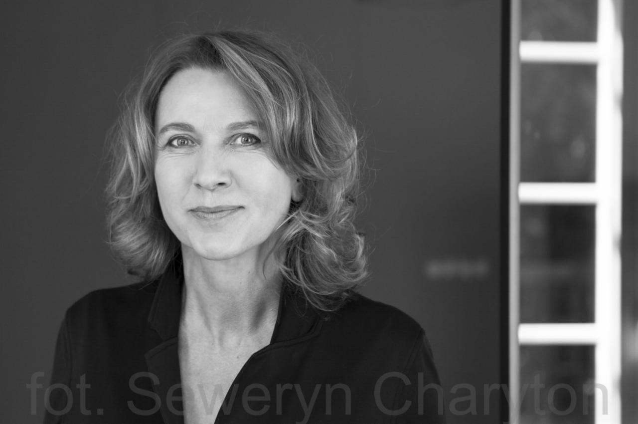 Portret kobiety o jasnych włosach, ubrana w ciemną marynarkę, na czarno-białym zdjęciu z widocznym podpisem fotografa.