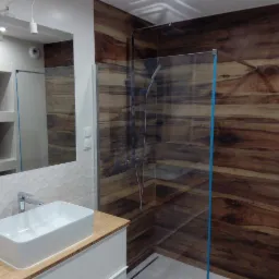 Nowoczesna łazienka z kabiną prysznicową typu walk-in, wykończona płytkami imitującymi drewno, białą umywalką nablatową i lustrem z oświetleniem punktowym.