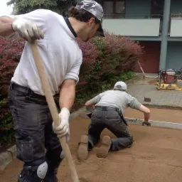 Dwóch mężczyzn w roboczych ubraniach wyrównuje piasek pod kostkę brukową, jeden z łopatą, drugi z poziomicą, na tle budynku z balkonami.