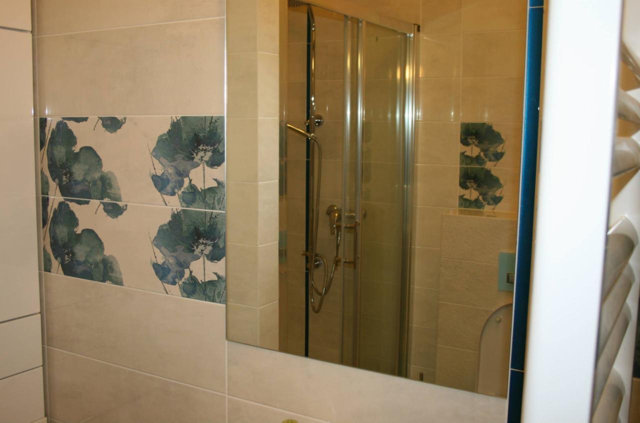 Beżowe płytki łazienkowe z motywem niebieskich kwiatów, odbicie kabiny prysznicowej w lustrze.