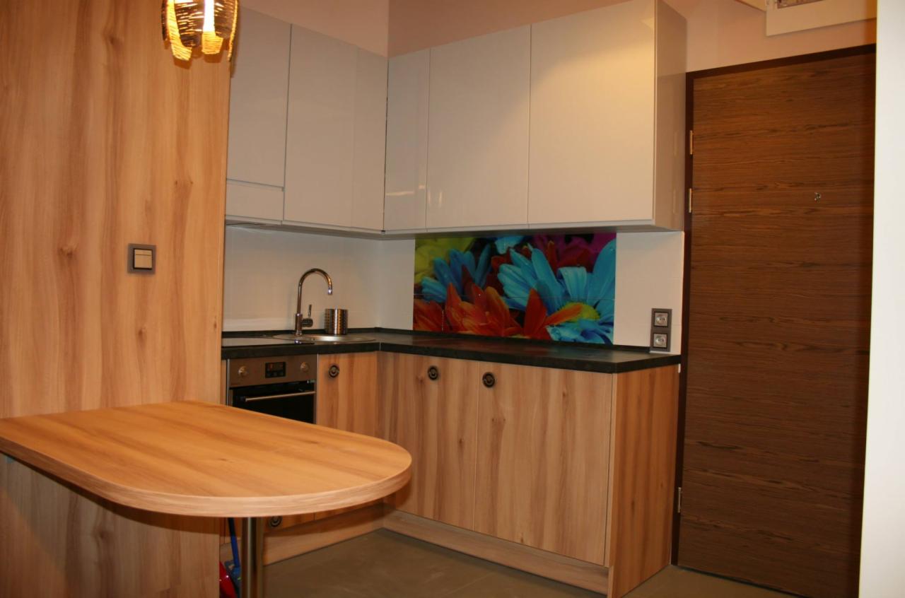 Jasne drewniane szafki kuchenne z czarnym blatem, kolorowy panel szklany z motywem kwiatowym nad blatem, okrągły stół z jasnego drewna i białe szafki wiszące.