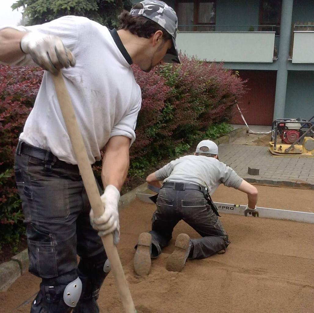 Dwóch mężczyzn w roboczych ubraniach wyrównuje piasek pod kostkę brukową, jeden z łopatą, drugi z poziomicą, na tle budynku z balkonami.