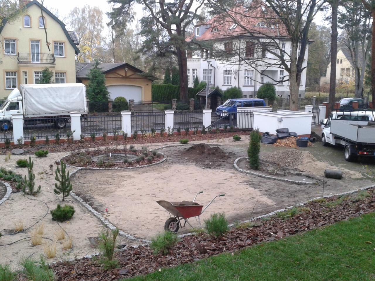 Aranżacja terenu przed posesją w trakcie realizacji: widoczne nasadzenia, alejki wysypane piaskiem, taczka, kopce ziemi, materiały budowlane, oraz samochody dostawcze na tle zabudowy jednorodzinnej.