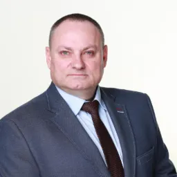 Portret mężczyzny w garniturze i krawacie na jasnym tle. Ujęcie od pasa w górę, z lekkim skrętem w lewo. Widoczna koszula, marynarka i krawat w brązowym odcieniu.