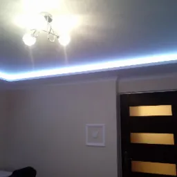 Pokój z zamontowanym oświetleniem LED podsufitowym, kremowe ściany, ciemne drzwi z poziomymi szybami i włącznik światła w białej ramce.