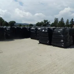 Rzędy czarnych, szczelnie owiniętych folią palet z towarem na placu składowym, widoczne drzewa i błękitne niebo w tle.