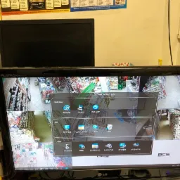 Ekran monitora LCD wyświetlający obraz z systemu kamer monitoringu w sklepie, z widocznym menu i logo BCS, ustawiony obok starszego monitora CRT i butelki spirytusu salicylowego.