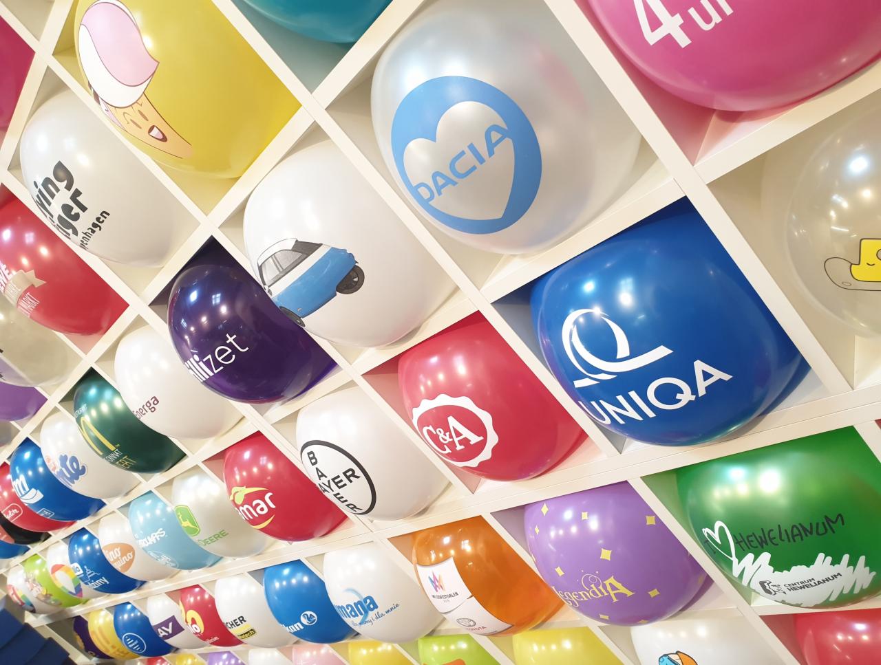Ściana z białych półek wypełnionych kolorowymi balonami z nadrukowanymi logotypami różnych firm, m.in. Dacia, Uniqua, Bayer, C&A, Energa, John Deere. Balony umieszczone w regularnych odstępach...