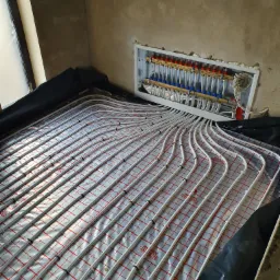 Rozłożona instalacja ogrzewania podłogowego z białych rurek na folii z czerwoną kratką, widoczny rozdzielacz w białej skrzynce na ścianie w surowym stanie.