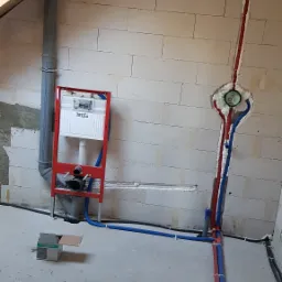 Instalacja podtynkowa TECE z czerwono-niebieskimi rurami PEX w surowym wnętrzu z bloków, widoczny również pion kanalizacyjny i pudełko Hansgrohe na podłodze.