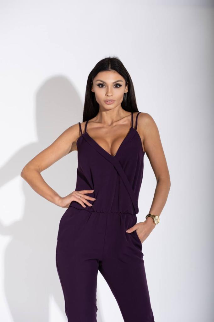 Kobieta w fioletowym kombinezonie z dekoltem w kształcie litery V, pozująca na białym tle, z ręką w kieszeni i złotym zegarkiem na nadgarstku.