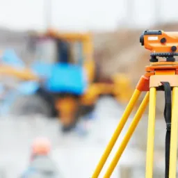 Pomarańczowy niwelator optyczny na żółtym statywie na placu budowy; w tle rozmyte maszyny budowlane i postać w pomarańczowym kasku.