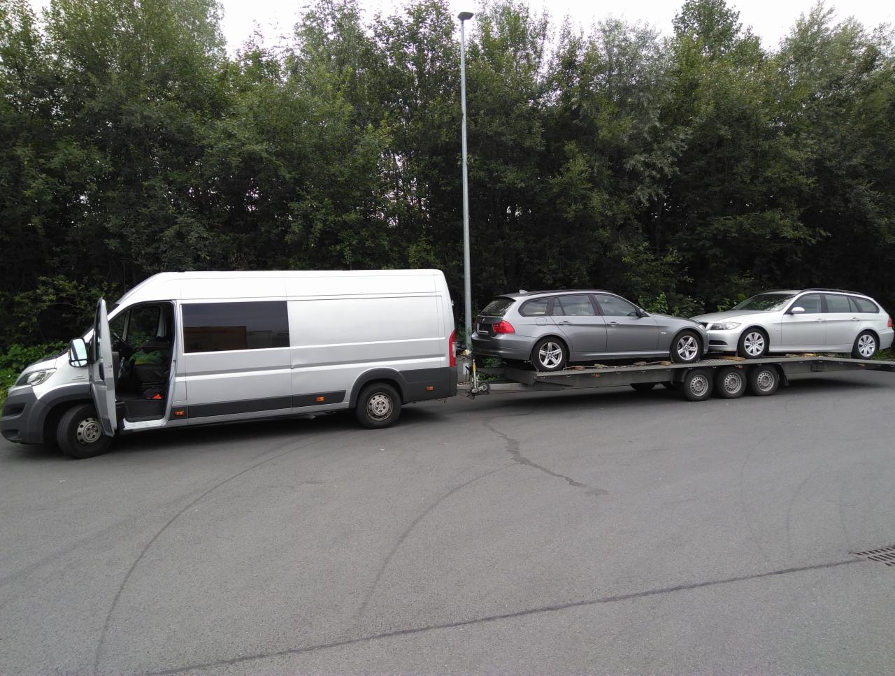 Srebrny bus z otwartymi drzwiami kierowcy ciągnie lawetę z dwoma srebrnymi samochodami typu kombi na tle zielonych drzew.