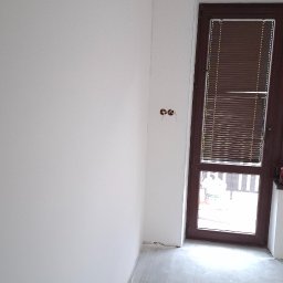 TermoGips Tomasz Buda - Białe ściany w pokoju po malowaniu, widoczne gniazdka elektryczne i brązowe drzwi z żaluzjami. Podłoga szara, surowa. Minimalistyczne wnętrze w trakcie wykańczania.