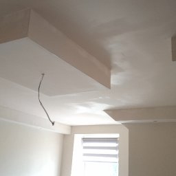 TermoGips Tomasz Buda - Białe, gładkie sufity z elementami dekoracyjnymi z płyty gipsowej, widok z dołu, kabel zwisający z sufitu, okno w tle. Jasne wnętrze po remoncie.