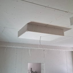 TermoGips Tomasz Buda - Sufit z płyt gipsowo-kartonowych z nietypową, wystającą konstrukcją w kształcie prostopadłościanu. Widoczne łączenia płyt i otwór drzwiowy w tle.