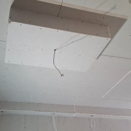 TermoGips Tomasz Buda - Sufit z płyt kartonowo-gipsowych z otworem rewizyjnym i wystającymi przewodami, widok z dołu, surowe wykończenie, drewniana framuga drzwi w prawym rogu.
