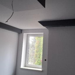 TermoGips Tomasz Buda - Szare ściany po malowaniu z czarnymi elementami dekoracyjnymi pod sufitem i oknem. Widoczne kable wystające z sufitu. W tle zieleń za oknem.