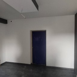 TermoGips Tomasz Buda - Wnętrze po malowaniu: białe ściany z ciemnymi akcentami na suficie i przy podłodze. Widoczne gniazdko elektryczne i niebieskie drzwi. Podłoga ciemna, surowa.