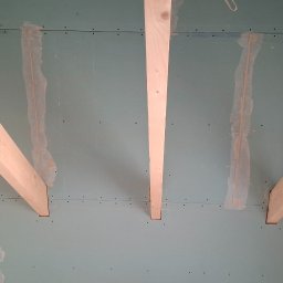 TermoGips Tomasz Buda - Montaż płyt gipsowo-kartonowych na suficie z widocznymi drewnianymi belkami i miejscami łączeń płyt. Widok z dołu, sufit w trakcie prac wykończeniowych.