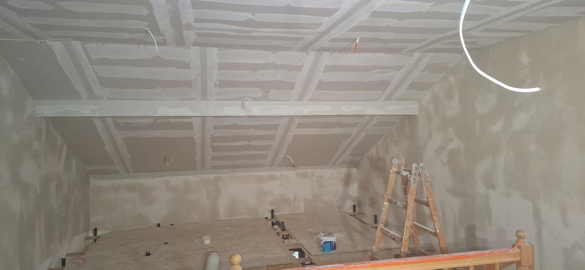 Poddasze w trakcie prac wykończeniowych: widoczne płyty gipsowo-kartonowe na skośnym suficie, częściowo wyszpachlowane ściany i drabina. W tle surowe deski podłogowe.