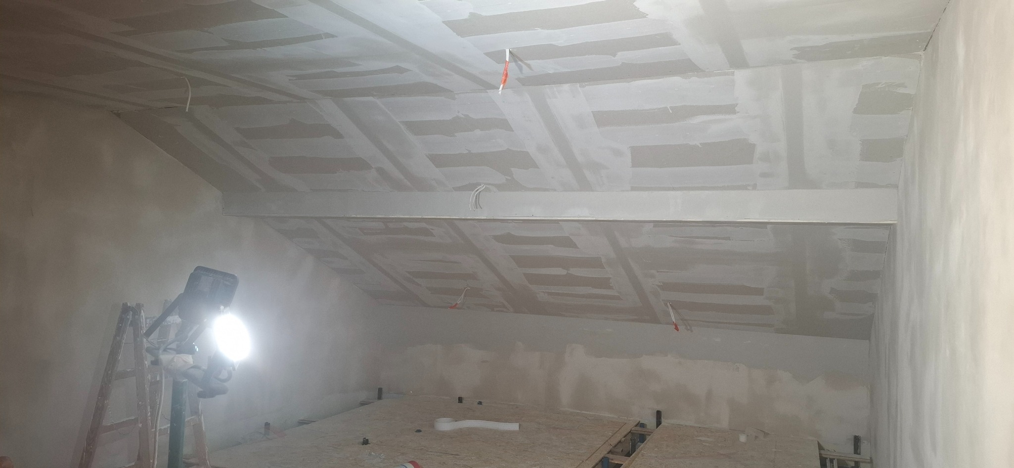 Skośny sufit poddasza po szpachlowaniu, widoczne łączenia płyt gipsowych, oświetlenie robocze na drabinie. W tle fragment podłogi z płyt OSB.