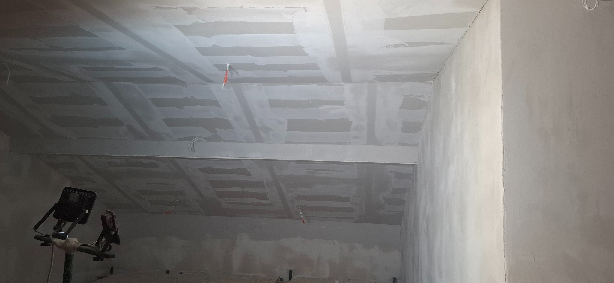 Skośny sufit poddasza po szpachlowaniu i montażu płyt gipsowych, widoczne kable i lampa budowlana. Ściany przygotowane do malowania.