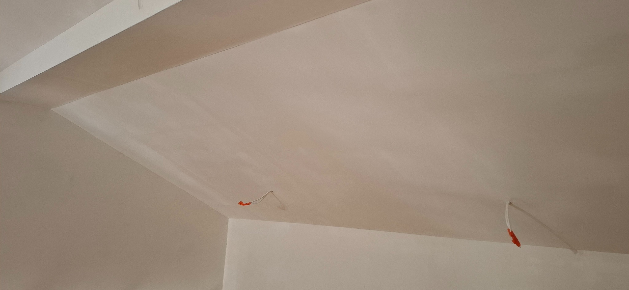 Gładka, jasna powierzchnia sufitu pod skosem, świeżo po szpachlowaniu, z widocznymi przewodami elektrycznymi wystającymi ze ściany. Minimalistyczne wykończenie.