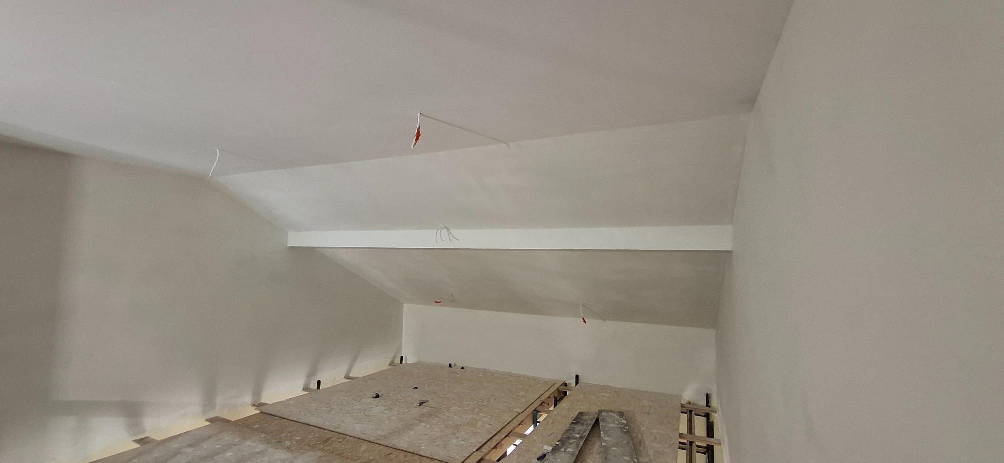 Poddasze po szpachlowaniu i malowaniu na biało, widoczne płyty OSB na podłodze i wystające kable. Ściany gładkie, jednolite, gotowe do dalszych prac wykończeniowych.