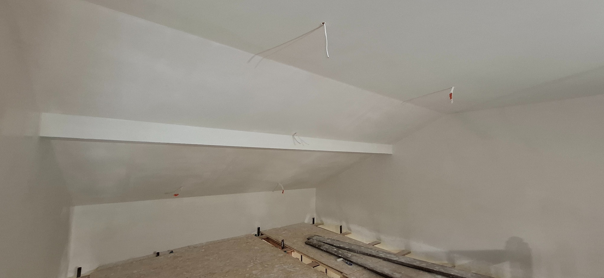 Poddasze po szpachlowaniu i malowaniu na biało, widoczne wystające kable i przygotowane podłoże pod podłogę. Skos dachu z belką.