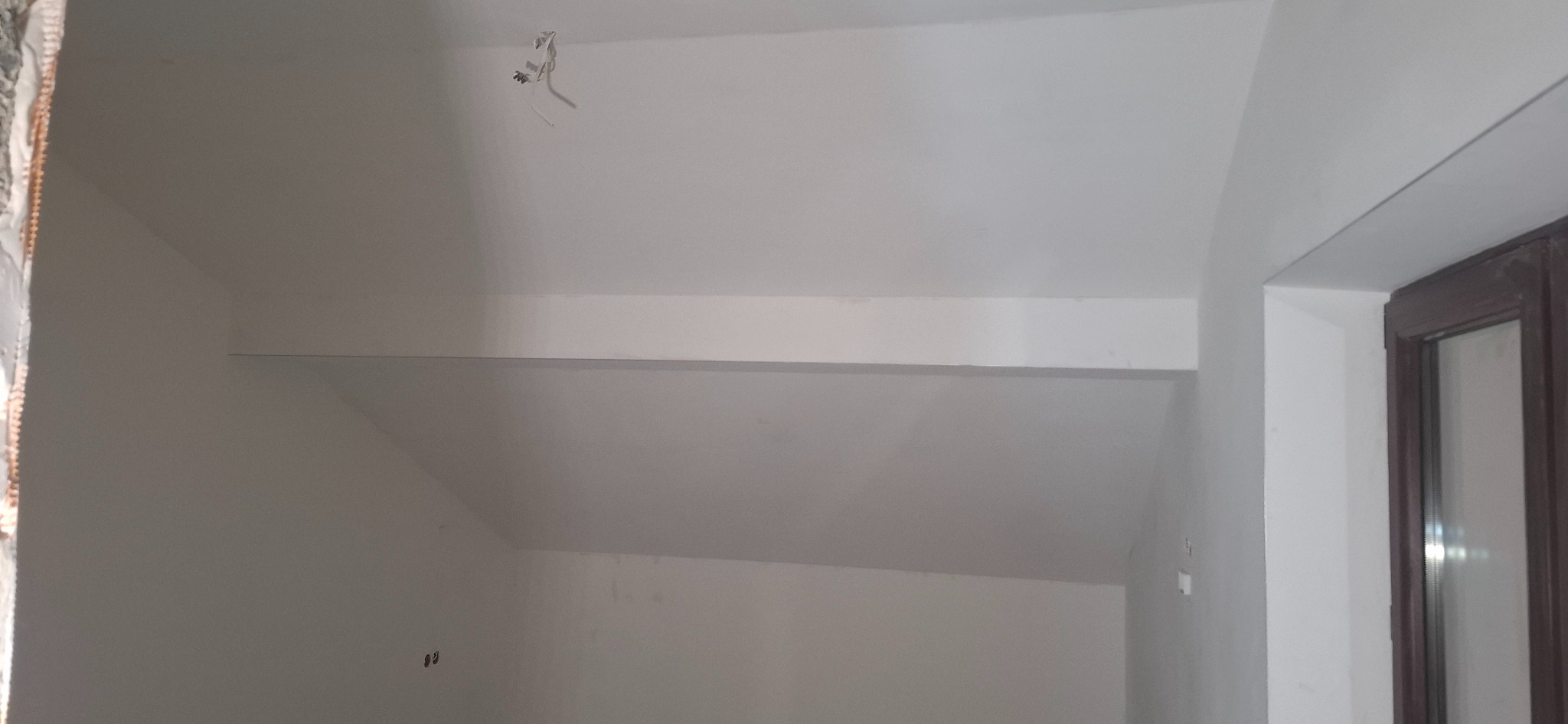 Wnętrze poddasza po remoncie: gładkie, białe ściany i sufit ze skosem, widoczne kable elektryczne, fragment okna. Minimalistyczny efekt wykończenia.