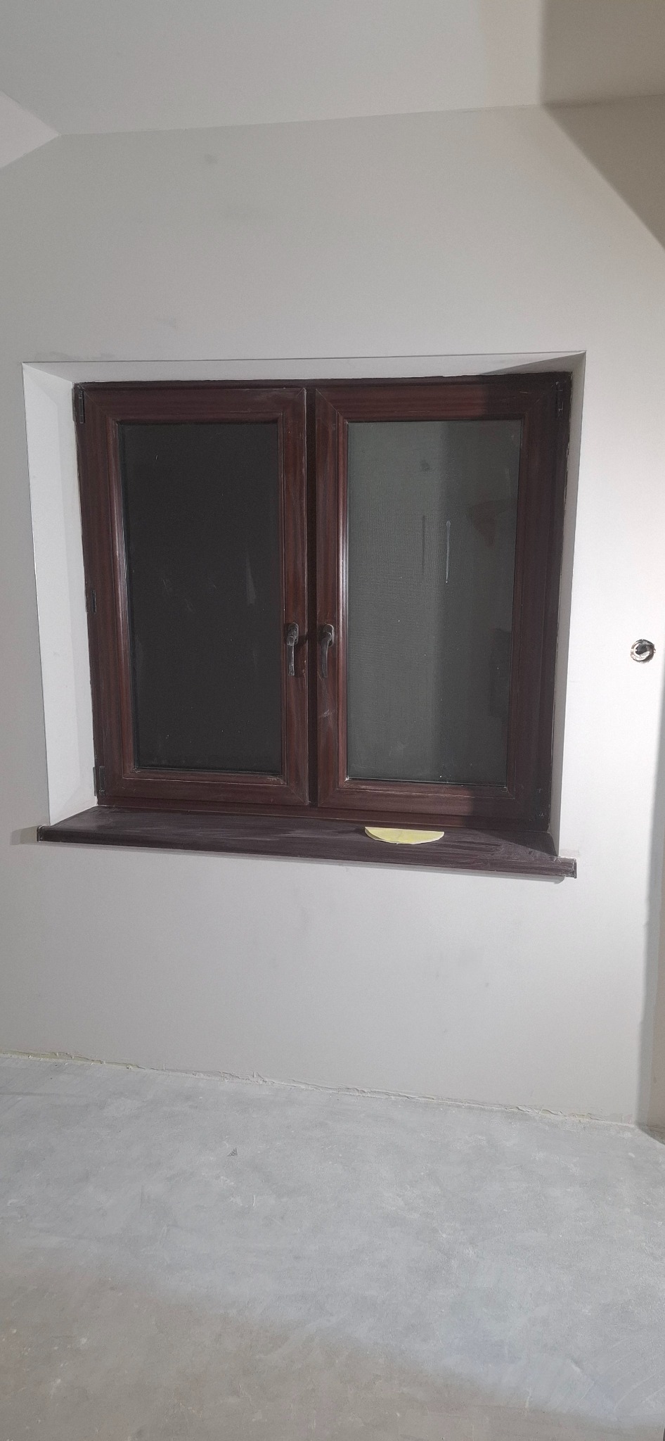 Białe ściany po szpachlowaniu z brązowym oknem i parapetem. Na parapecie leży żółta szpachelka. Widoczna surowa podłoga. Świeżo wykończone wnętrze w trakcie prac.