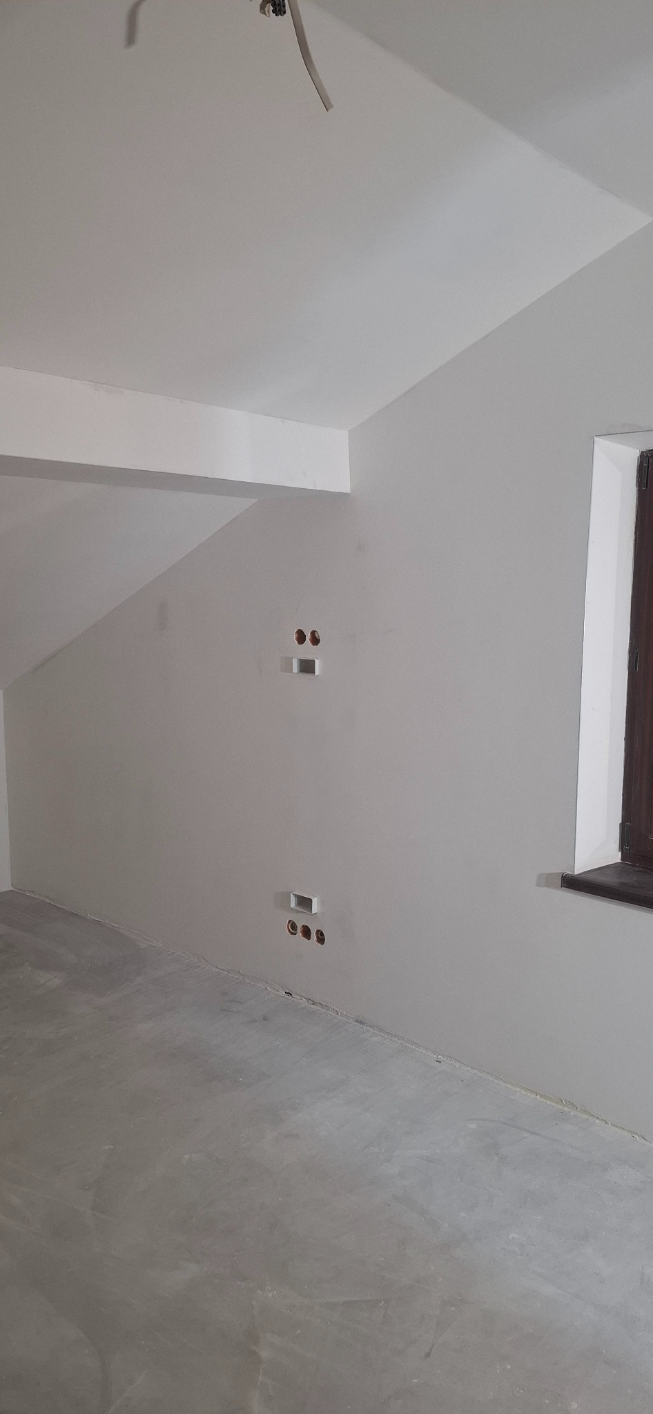 Wnętrze poddasza w Żarnówce po szpachlowaniu, z widocznymi otworami na gniazdka elektryczne i oknem z ciemną ramą. Ściany i sufit pomalowane na biało.