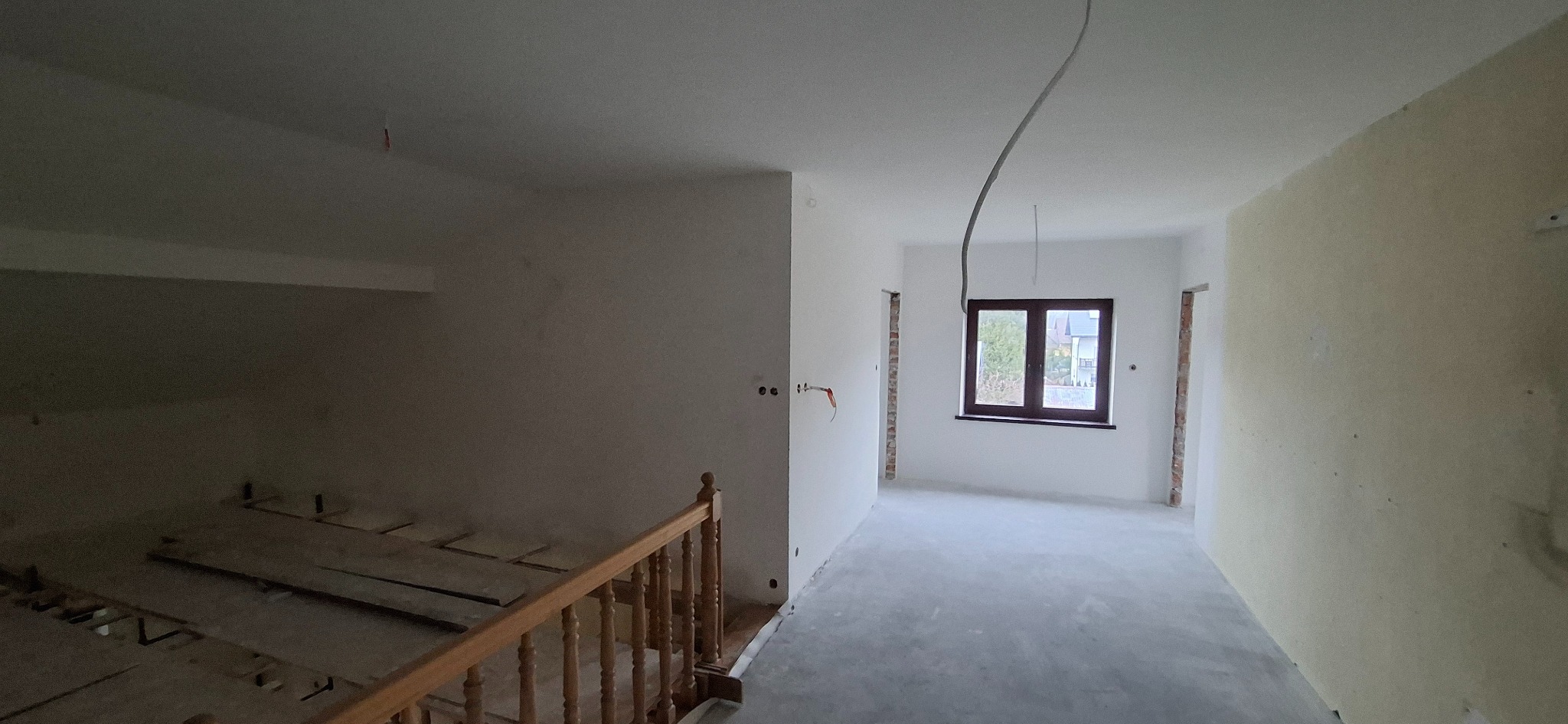 Wnętrze w trakcie remontu: gładkie, białe ściany, okno z widokiem na zieleń, drewniane schody. Widoczne ślady po instalacjach elektrycznych i brakujące panele podłogowe.