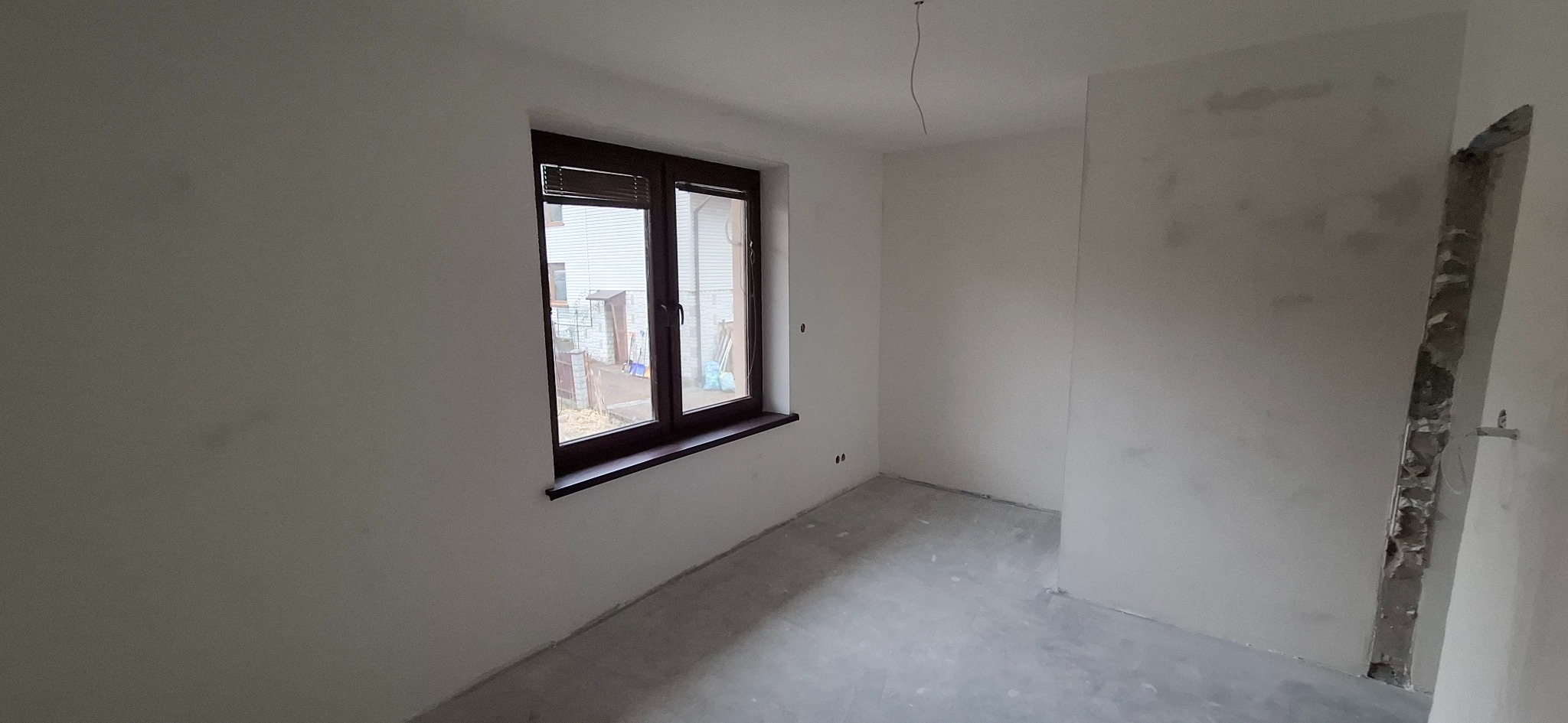 Pokój po szpachlowaniu ścian, z widocznym oknem z ciemną ramą i surowym betonem na podłodze. Ściany przygotowane do dalszych prac wykończeniowych.