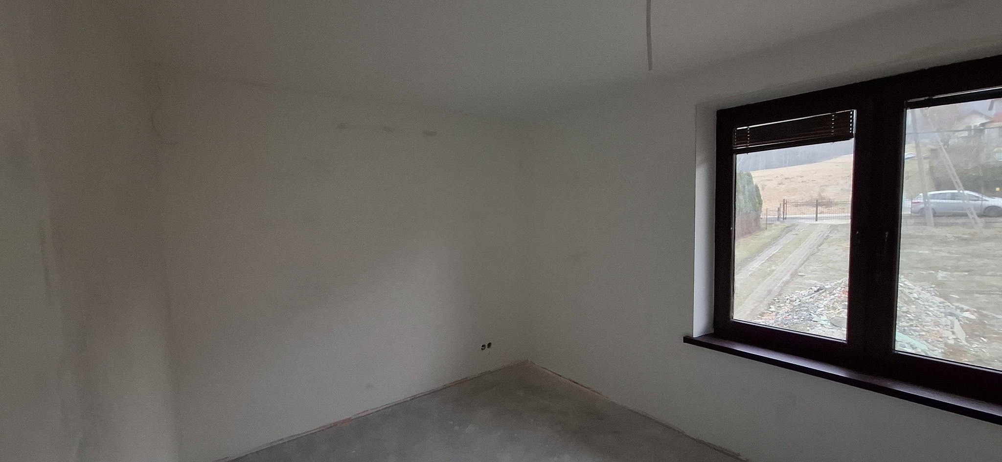 Jasne wnętrze po szpachlowaniu, z widocznymi gładkimi ścianami i oknem z brązową ramą. Świeżo wykończone ściany gotowe do malowania. Widok na zewnątrz przez okno.