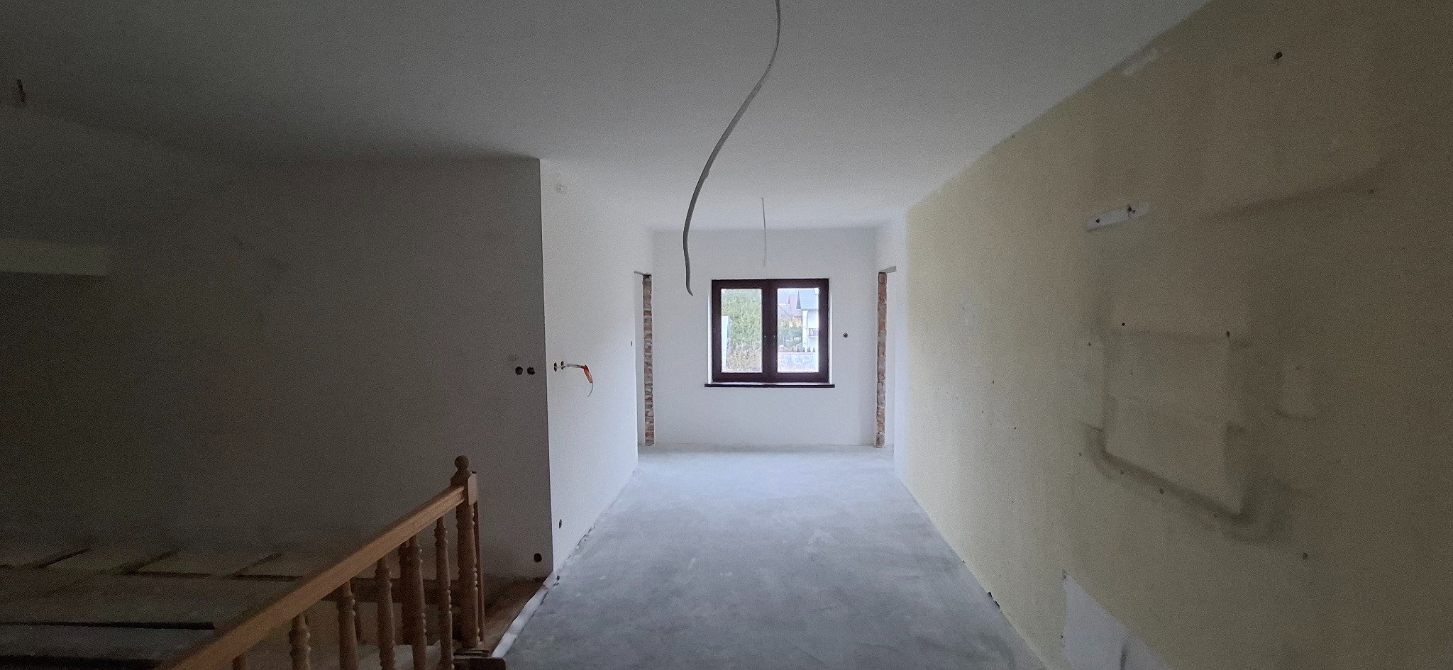 Wnętrze w trakcie prac wykończeniowych: surowe ściany, widoczne przewody, okno z widokiem na zieleń. Częściowo wykończona podłoga i fragment drewnianej balustrady schodów.