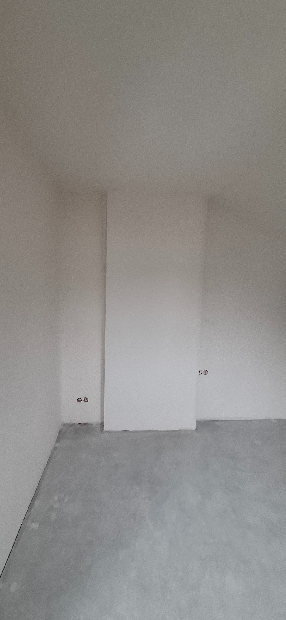Wnętrze pokoju po szpachlowaniu ścian i wylewce podłogi. Widoczne gniazdka elektryczne. Ściany i sufit w kolorze białym, podłoga szara.