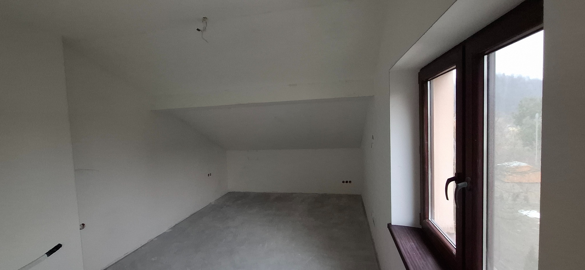 Poddasze po szpachlowaniu i malowaniu na biało, widoczne gładkie ściany, okno z brązową ramą i widok na zewnątrz. Przygotowane do dalszych prac wykończeniowych.