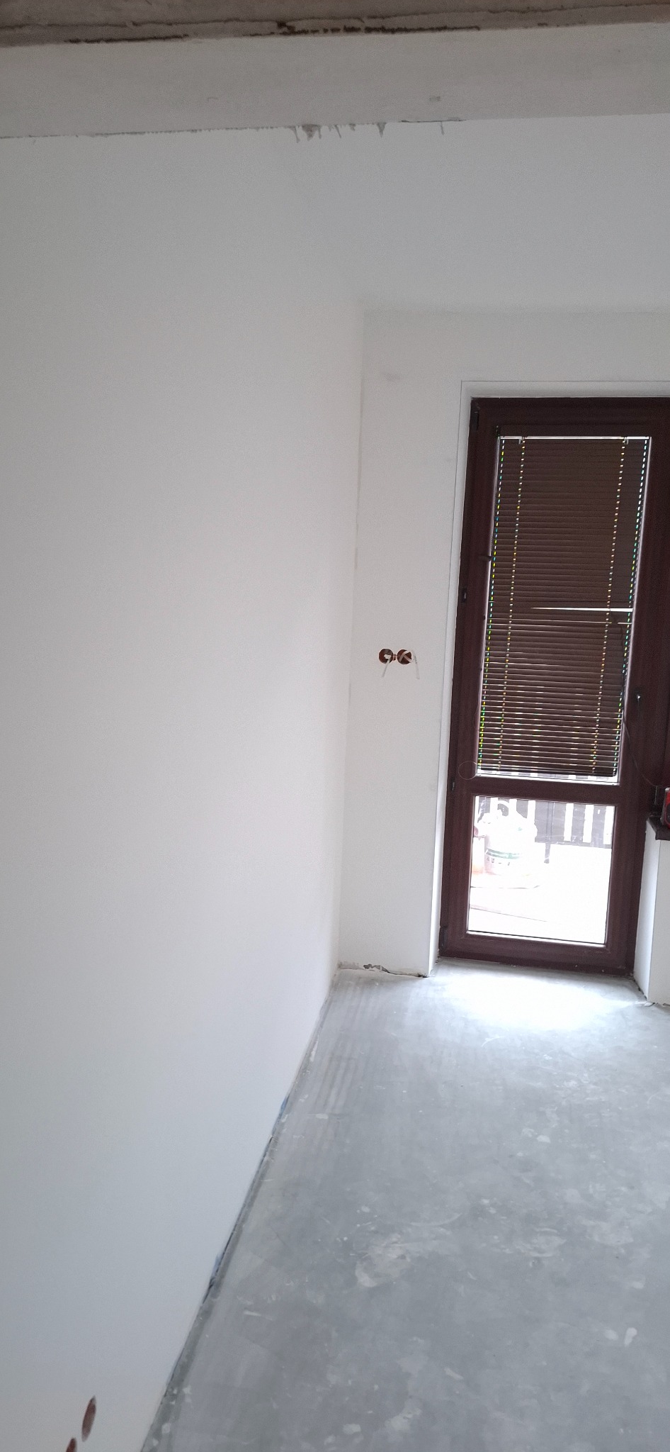 Białe ściany w pokoju po malowaniu, widoczne gniazdka elektryczne i brązowe drzwi z żaluzjami. Podłoga szara, surowa. Minimalistyczne wnętrze w trakcie wykańczania.