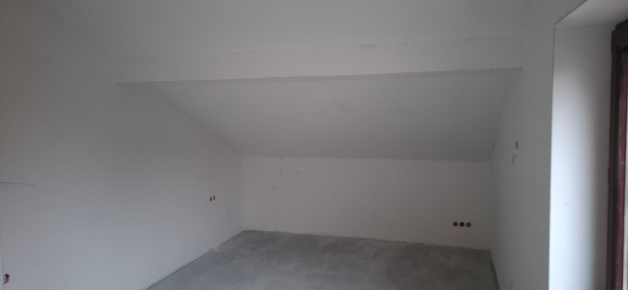 Wnętrze poddasza po szpachlowaniu i malowaniu na biało, widoczne gniazdka elektryczne i okno po prawej stronie. Ściany i sufit gładkie, gotowe do dalszych prac.