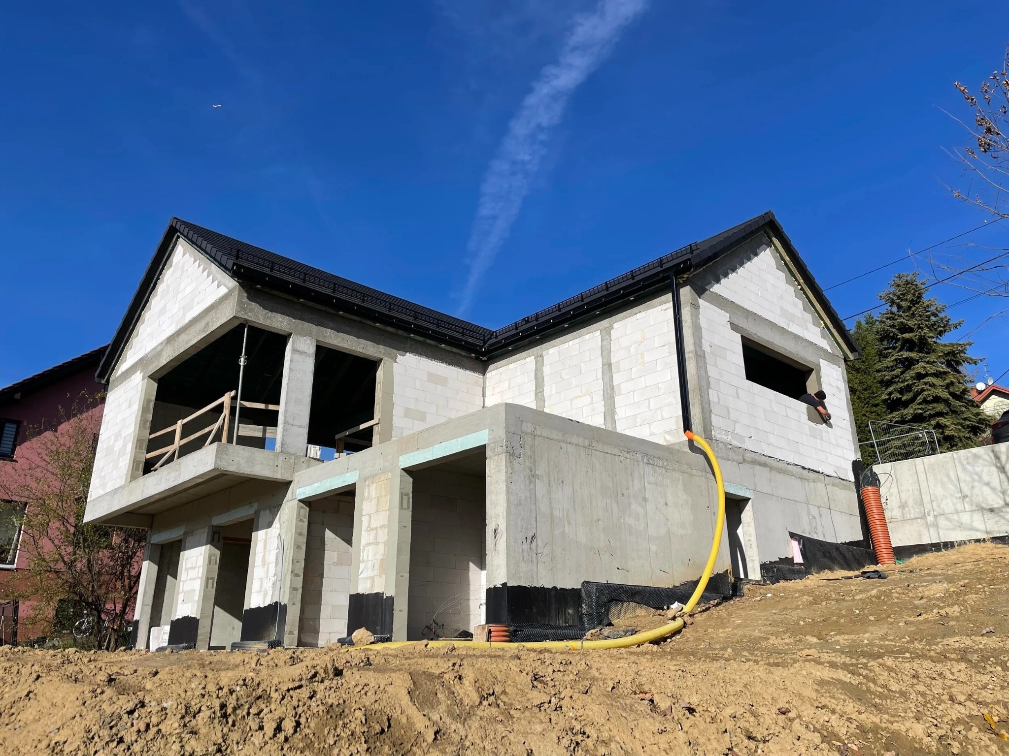 Budowa domu jednorodzinnego w Wieliczce w stanie surowym otwartym, widok z dołu na tle błękitnego nieba z smugami kondensacyjnymi. Żółty wąż na ziemi.