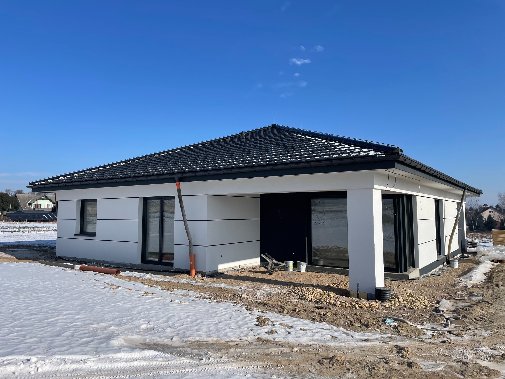 Nowoczesny, parterowy dom jednorodzinny z czarnym dachem i białymi ścianami w zimowej scenerii. Widoczne detale wykończeniowe i teren wokół budynku w trakcie prac.