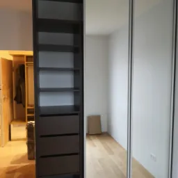 Nowoczesna szafa wnękowa z przesuwnymi drzwiami lustrzanymi i brązowym regałem z półkami i szufladami, widok na wnętrze pokoju z drewnianą podłogą.