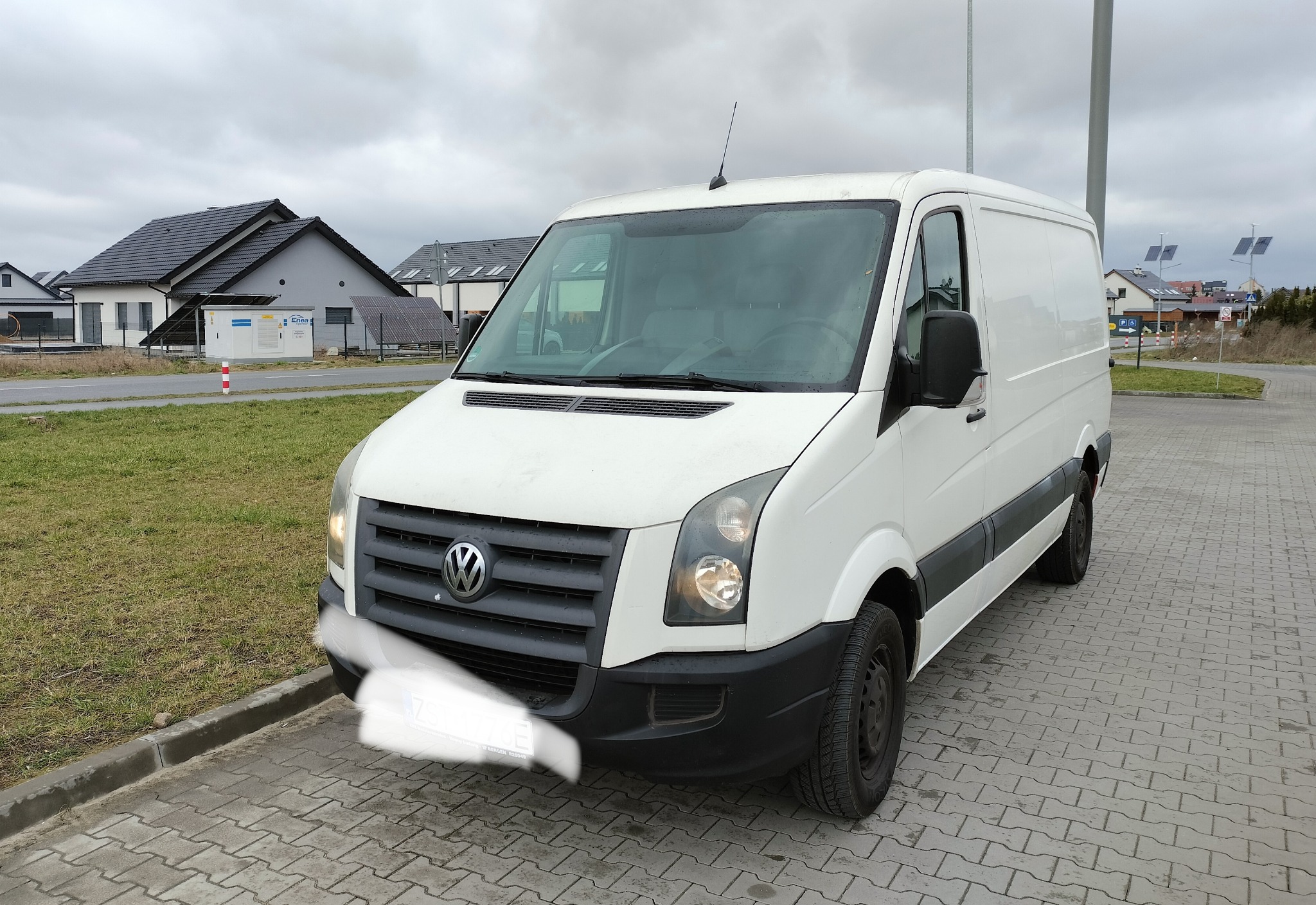 Biały van Volkswagen Crafter na kostce brukowej, z widocznymi domami jednorodzinnymi w tle. Światła włączone, pochmurne niebo.