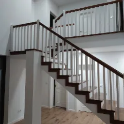 Nowoczesne schody drewniane z białą balustradą i drewnianymi stopniami, prowadzące na antresolę w przestronnym, jasnym wnętrzu.