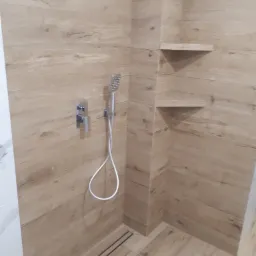 Nowoczesna kabina prysznicowa wykończona płytkami imitującymi drewno, z chromowaną armaturą i odpływem liniowym, uzupełniona o dwie półki wnękowe.