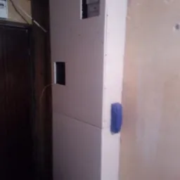 Konstrukcja z płyt gipsowo-kartonowych z widocznymi otworami na instalacje, niezagruntowana, z niebieską gąbką przytwierdzoną do boku, na tle ściany wymagającej odświeżenia.